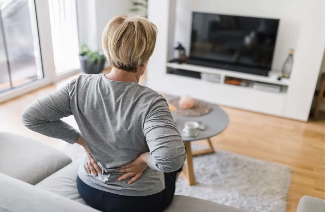 Older woman seen from behind on a couch, holding both hands to her lower back