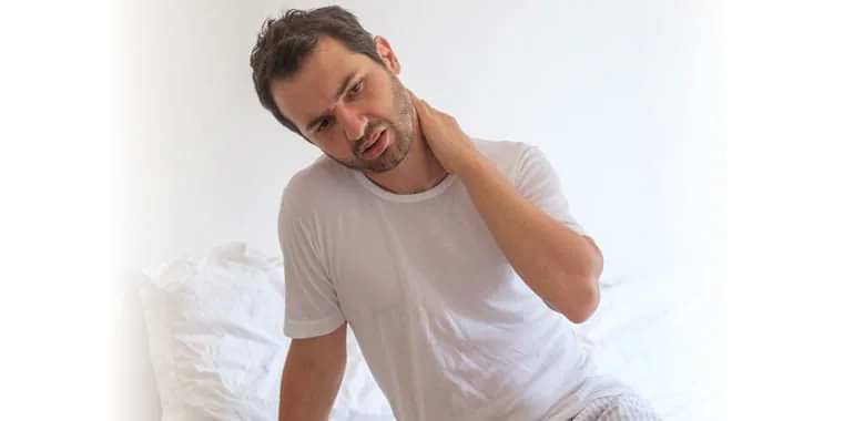 Man on a bed with a pained expression, holding the back of his neck