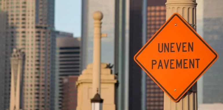 Orange road sign reading Uneven Pavement in front of city buildings