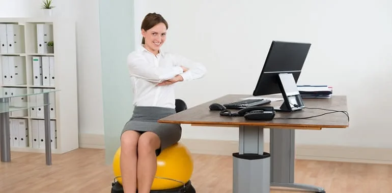 Smiling woman stretching her arms across her chest while seated on a yellow exercise ball at her desk