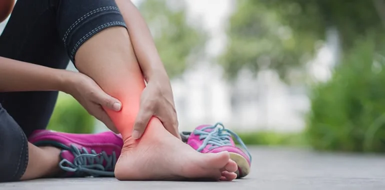 Person sitting outdoors in sneakers gripping their ankle, with a red glow marking pain