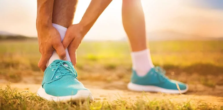 Runner in teal sneakers gripping an injured ankle on a dirt trail at sunset