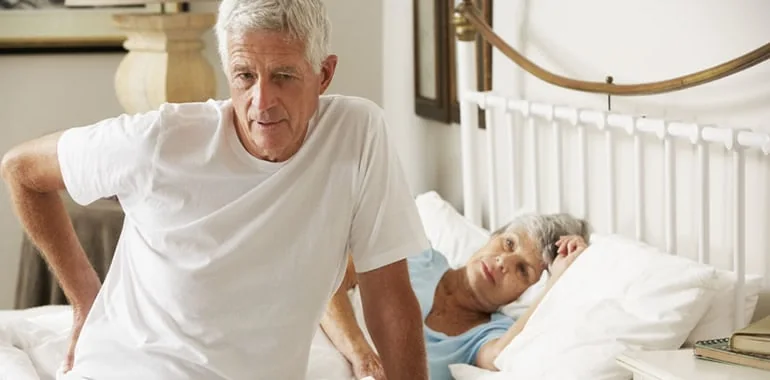 Older man sitting up in bed with his hand on his lower back while his wife lies beside him